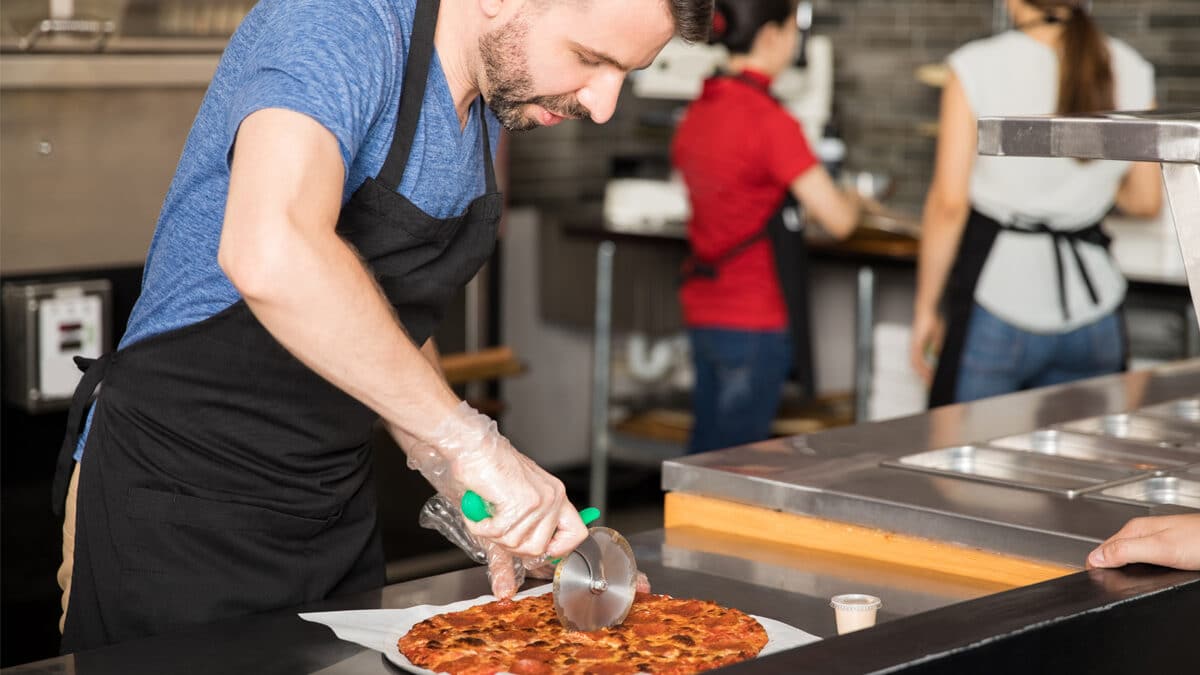 Man in blue shirt and apron cutting a pizza for delivery.