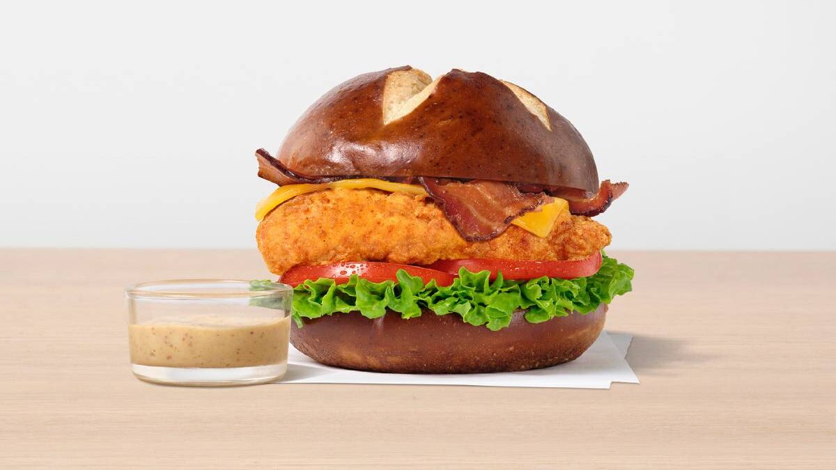 The sandwich features a toasted, buttery pretzel bun, lettuce, sliced tomato, cheddar cheese, strips of applewood smoked bacon and a side of Creamy Dijon Mustard Sauce.