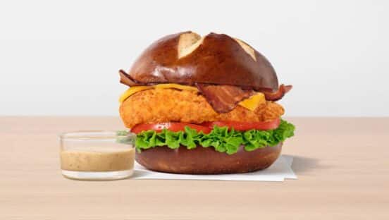 The sandwich features a toasted, buttery pretzel bun, lettuce, sliced tomato, cheddar cheese, strips of applewood smoked bacon and a side of Creamy Dijon Mustard Sauce.