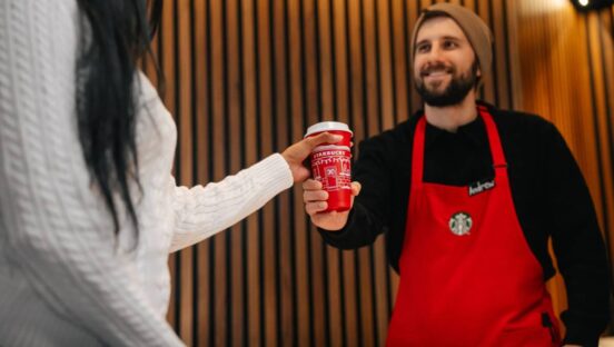 Starbucks employee hands over red cup in red apron to customer.